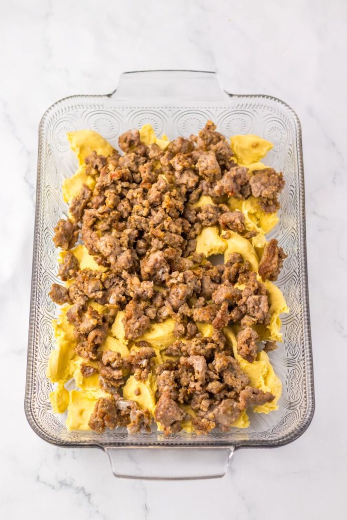 Raw biscuit dough topped with cooked sausage in glass casserole dish.