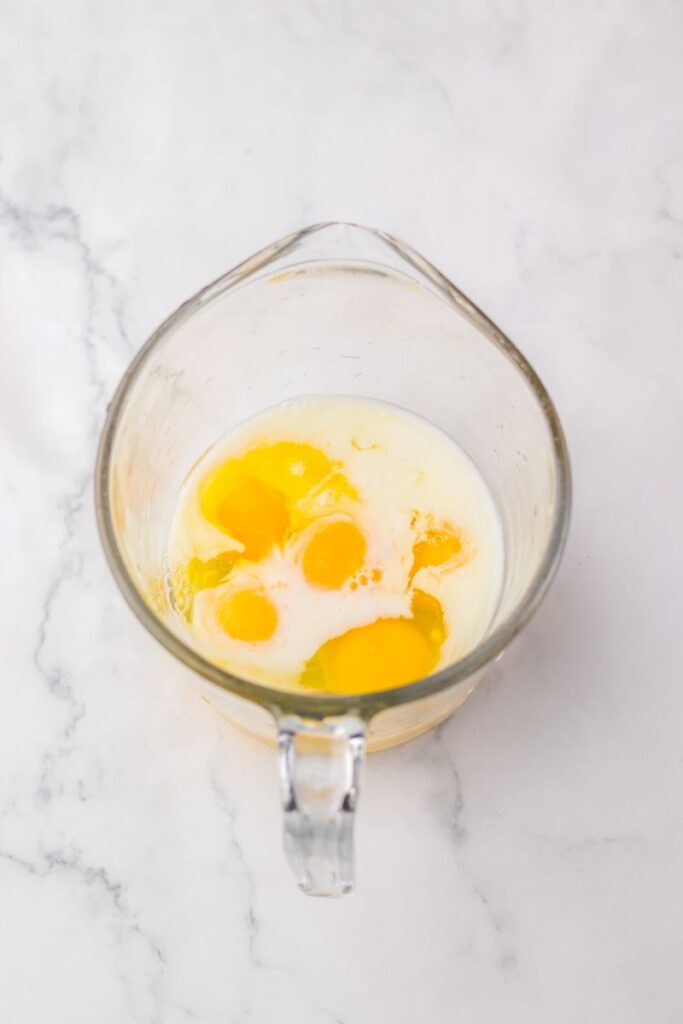 Overhead view of clear glass measuring cup with milk and raw eggs.