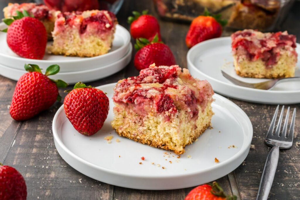Close up of one piece of strawberry coffee cake on white plate next to whole strawberries.