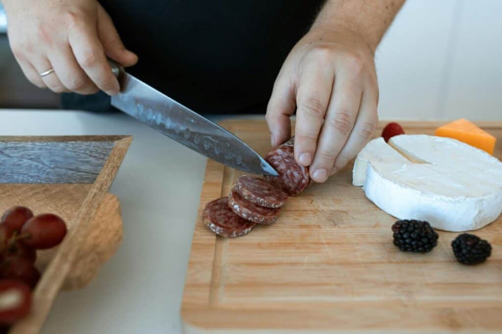 Hands chopping salami on wooden cutting board.