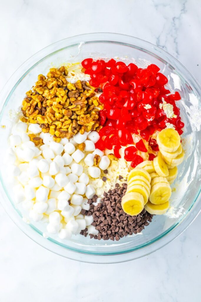 Overhead view of chopped walnuts, chopped red cherries, sliced bananas, mini chocolate chips, and mini marshmallows in glass bowl.