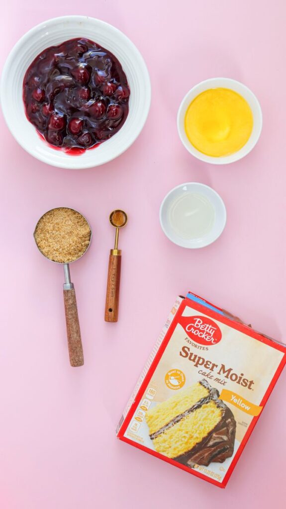 Overhead view of ingredients for cherry dump cake in white bowls on pink surface.
