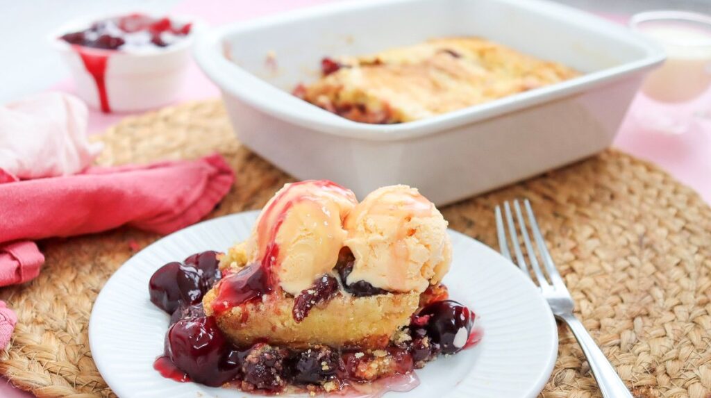 Side view of cherry dump cake topped with ice cream on white plate.
