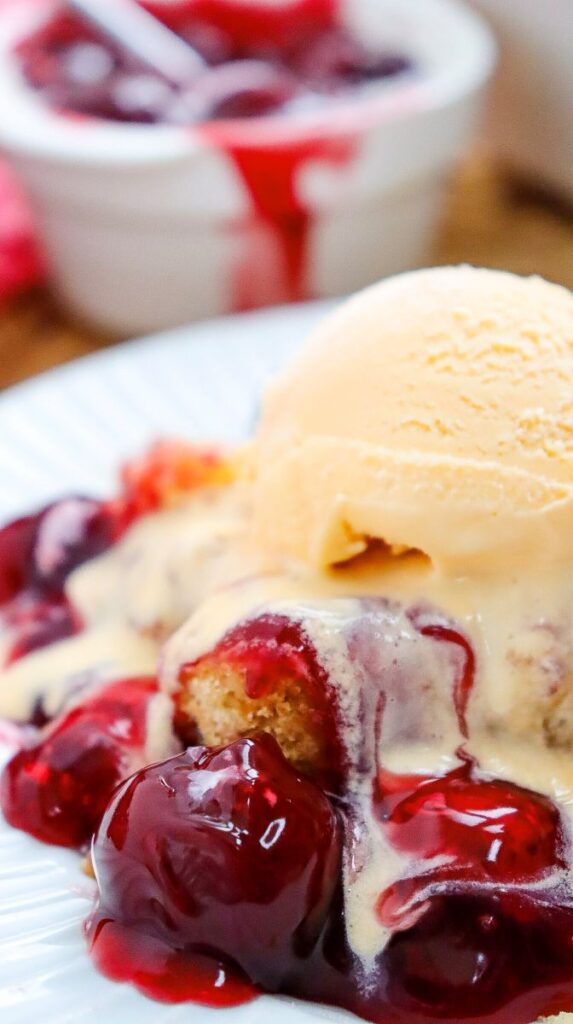 Close up of one serving of cherry dump cake topped with ice cream on white plate.