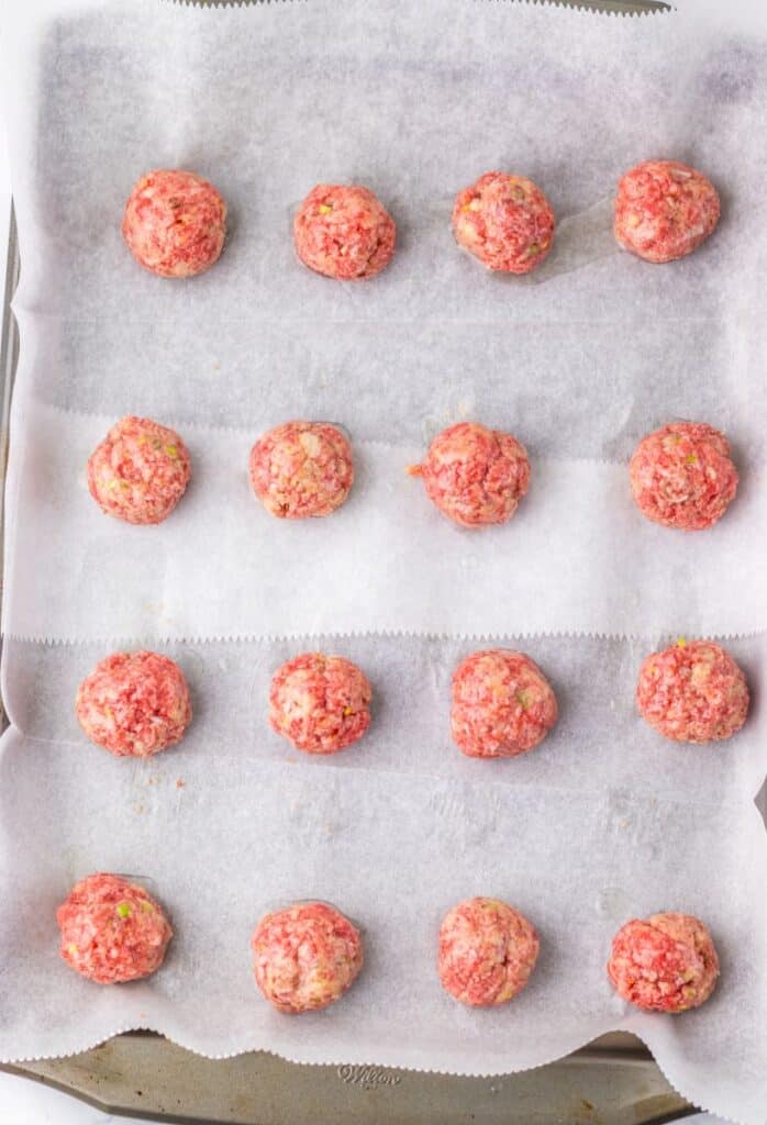 16 raw ground beef meatballs on parchment-paper lined baking sheet.