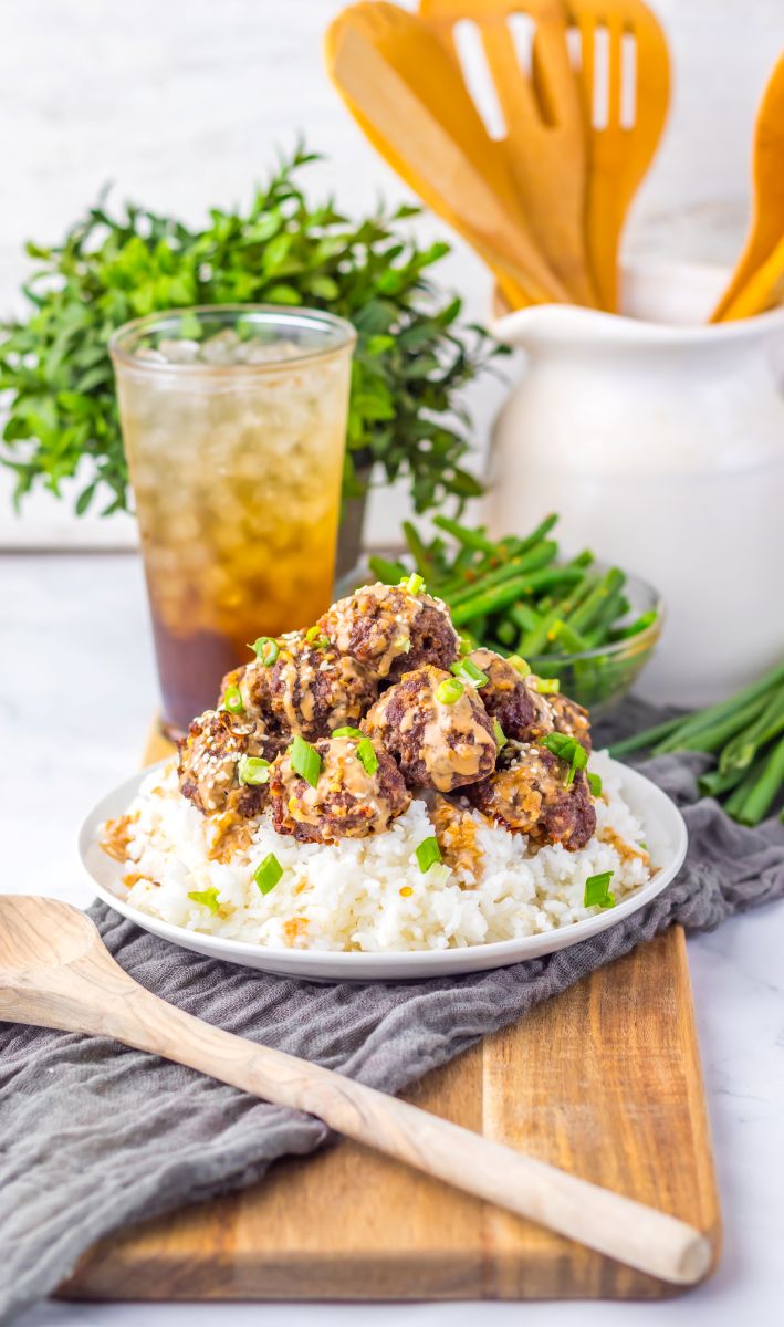 Side view of meatballs over white rice on white plate with dark brown drink in the background.