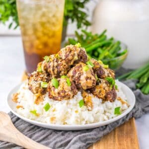 Meatballs over white rice on white plate with dark brown drink in the background.