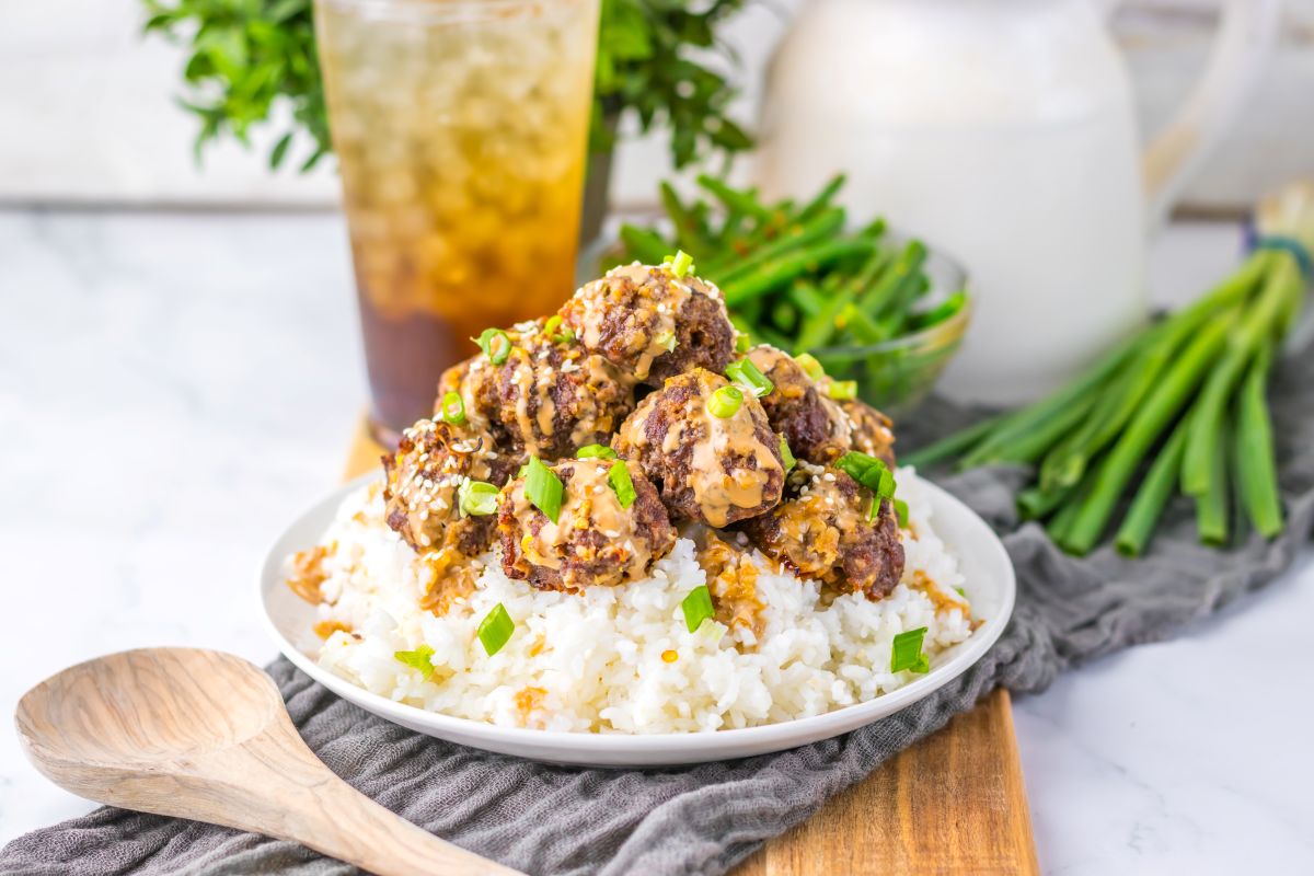 Meatballs over white rice on white plate with dark brown drink in the background.