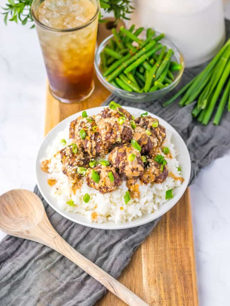 Meatballs over white rice on white plate with dark brown drink in the background.