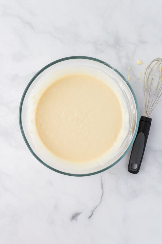 Overhead view of pancake batter in glass bowl next to whisk on white background.