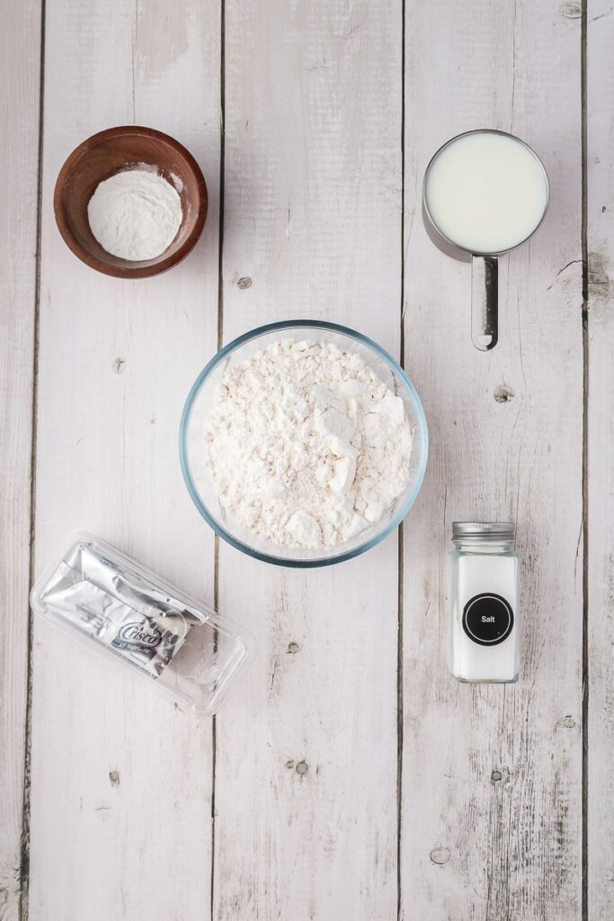 Overhead view of ingredients for baking powder biscuits on white wooden surface.