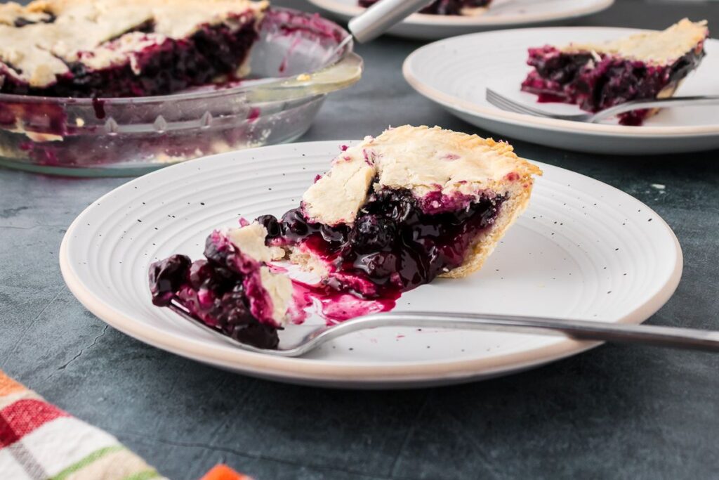 Close up of one bite of blueberry pie on fork white plate sitting next to the rest of the slice.