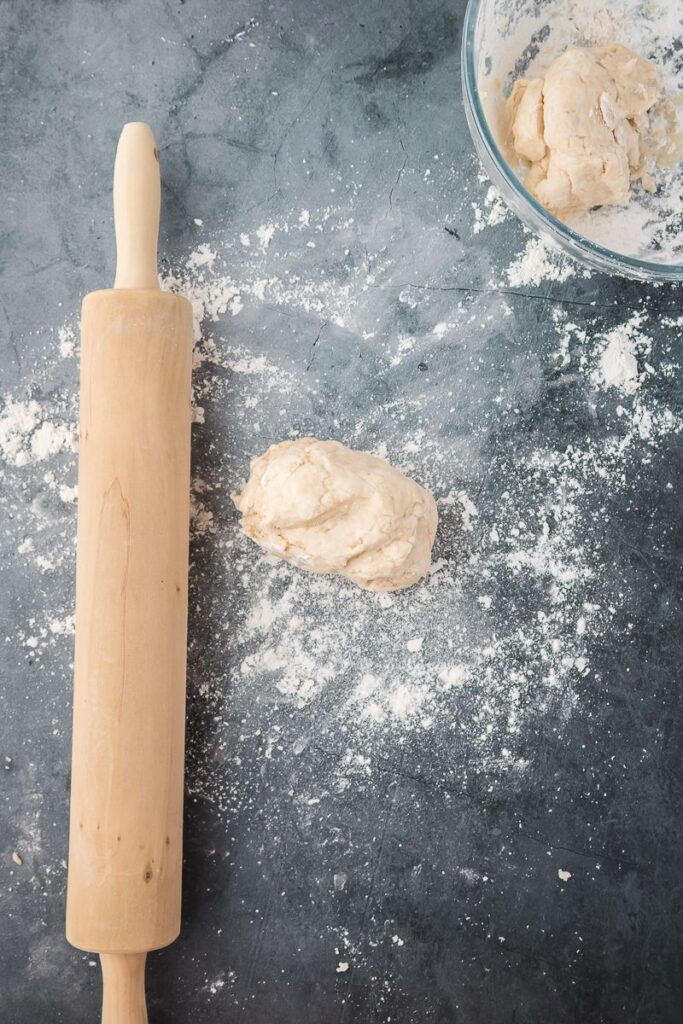 Wooden rolling pin next to ball of dough on floured dark surface.