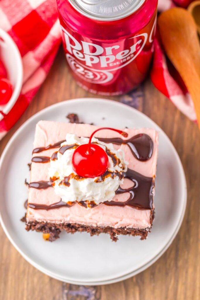 Close up of one slice of chocolate cake with pink frosting topped with whipped cream, a cherry, and chocolate drizzle.