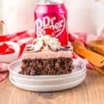 Side view of one slice of chocolate cake with pink frosting topped with whipped cream, a cherry, and chocolate drizzle.