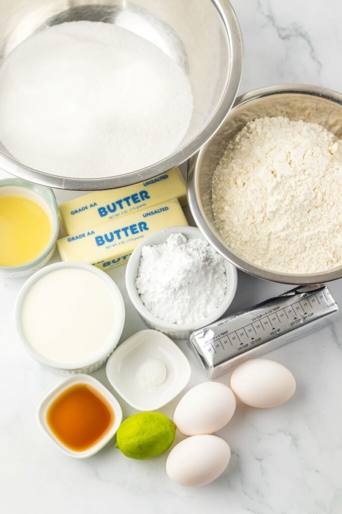 Baking ingredients on a white surface, including bowls of sugar and flour, sticks of butter, eggs, vanilla extract, milk, lime, powdered sugar, oil, and baking powder.
