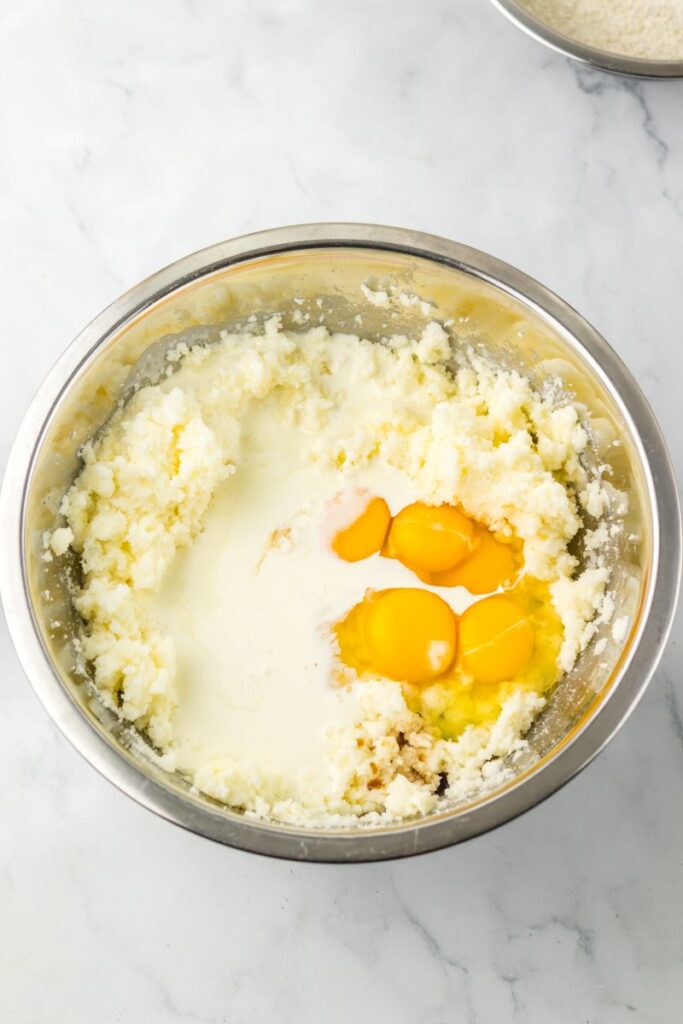A metal mixing bowl containing creamed butter and sugar, with three raw eggs and some milk added on top, ready to be mixed, on a white marble surface.