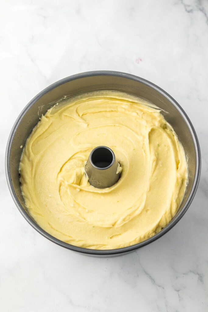 A round bundt cake pan filled with smooth, yellow cake batter, ready to be baked, sits on a white marble surface.