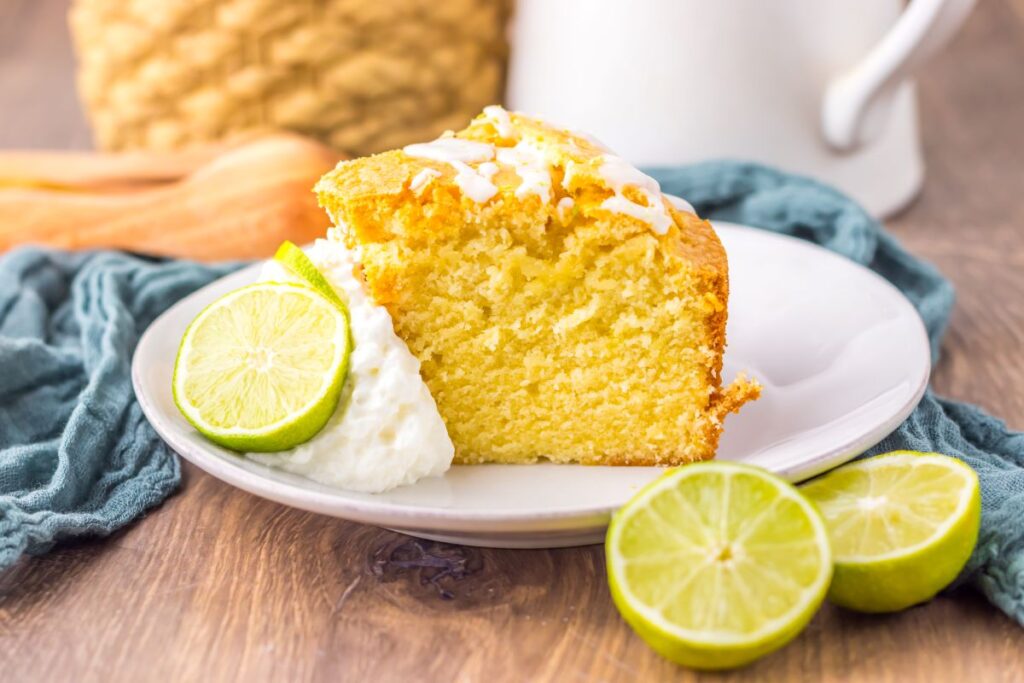 A slice of yellow cake with white icing is served on a plate with whipped cream and lime slices, set on a wooden table with a blue cloth and blurred background items.