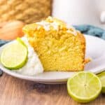 A slice of yellow cake with white icing is served on a plate with whipped cream and lime slices, set on a wooden table with a blue cloth and blurred background items.