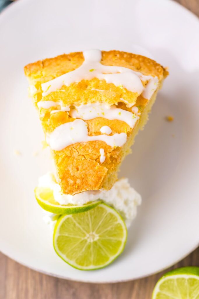 A slice of yellow cake with white icing on top, served on a white plate with a dollop of whipped cream and two lime slices on the side.