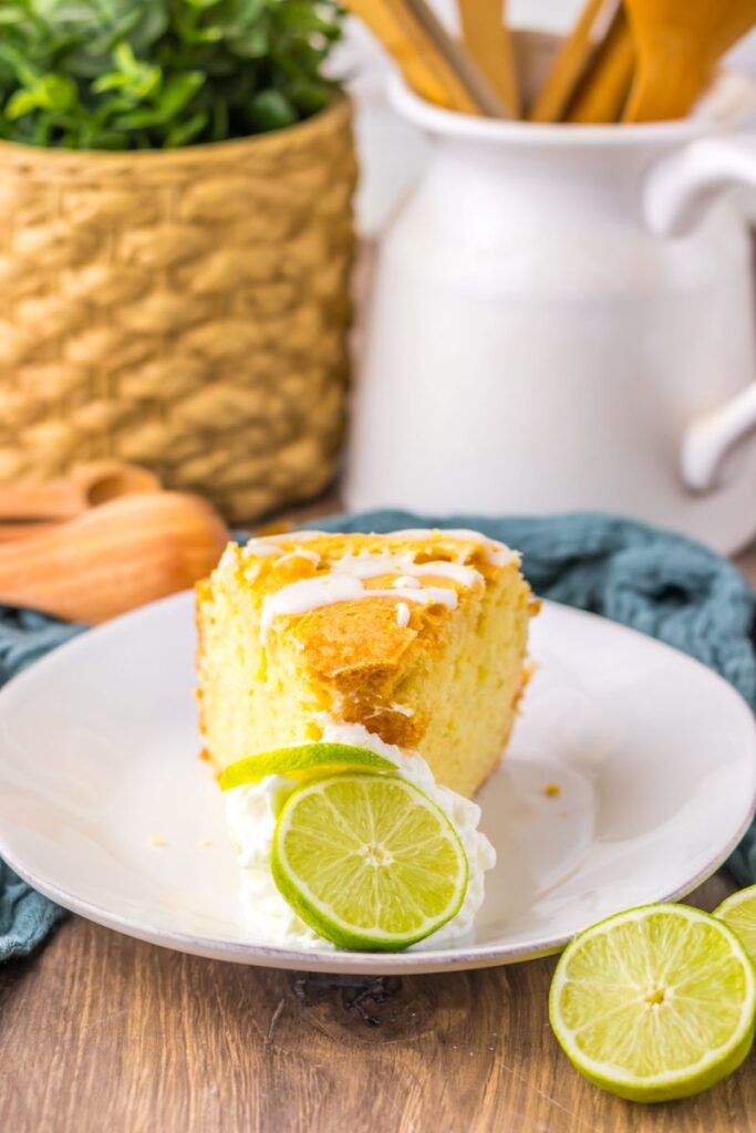 A slice of lime cake with white icing sits on a white plate, topped with whipped cream and a lime slice. A cut lime, woven basket, and kitchen utensils are in the blurred background.