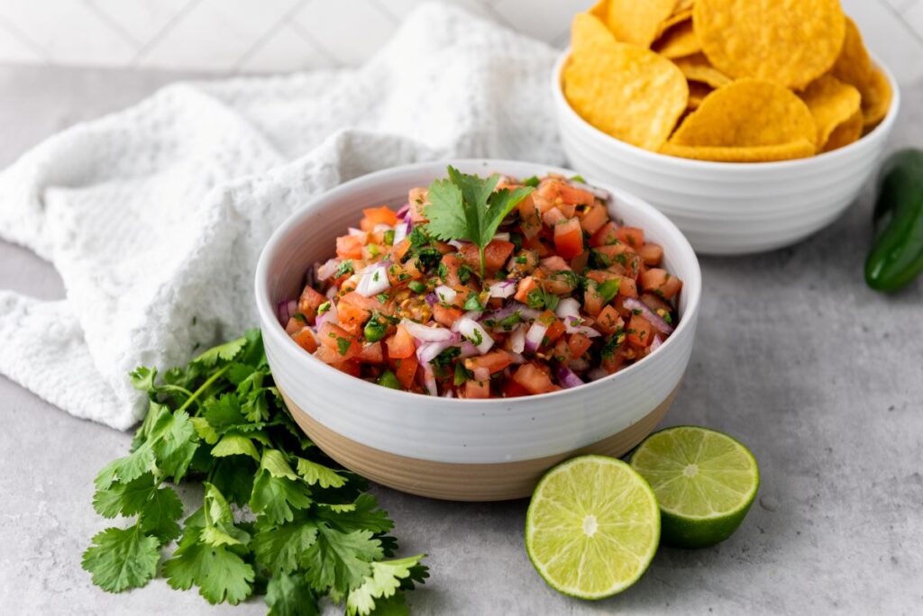 Side view of white bowl with salsa in front of cilantro and two halves of a lime.