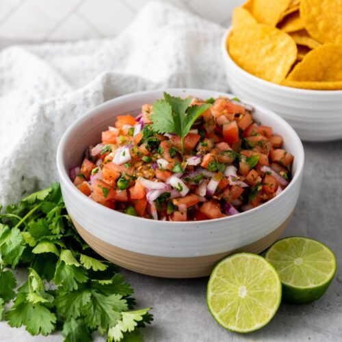 Side view of white bowl with salsa in front of cilantro and two halves of a lime.