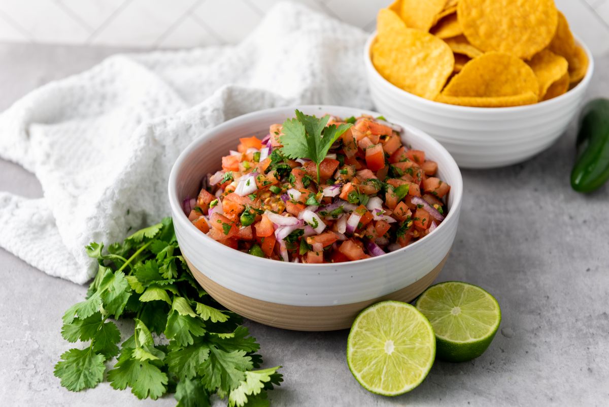Side view of white bowl with salsa in front of cilantro and two halves of a lime.