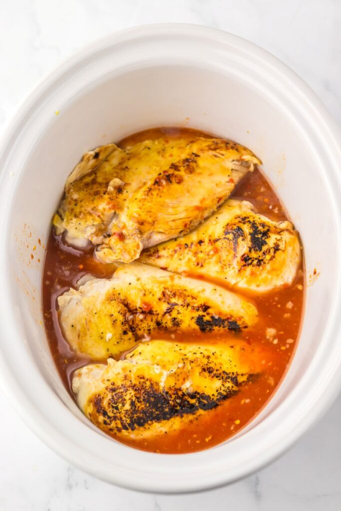 Overhead view of seared chicken with red sauce in white slow cooker.