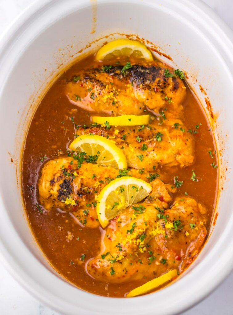 Overhead view of cooked chicken with lemons and reddish brown sauce in white slow cooker.