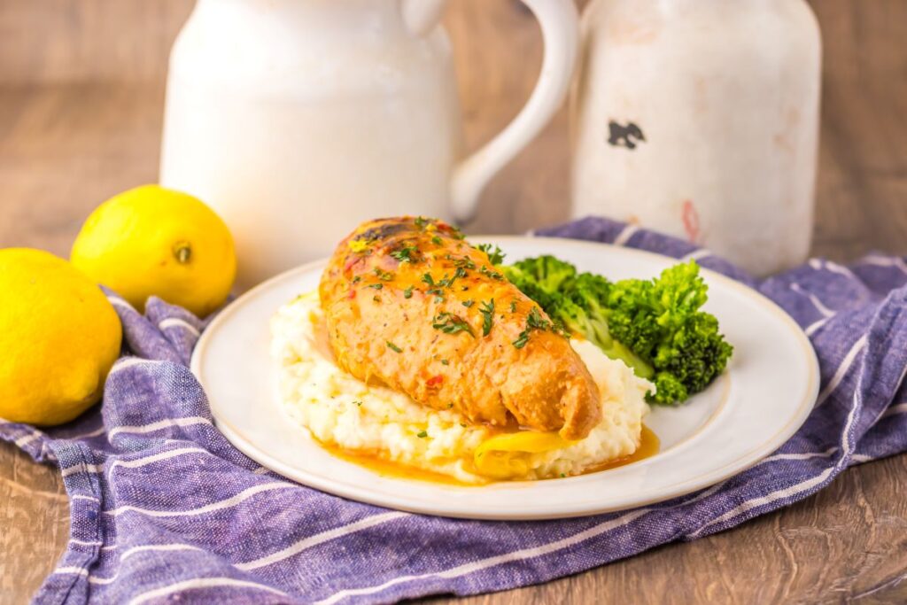 Side view of cooked chicken breast on mashed potatoes next to cooked broccoli on white plate.