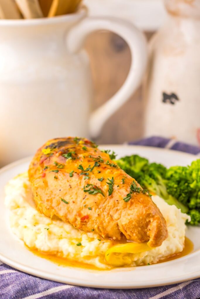 Close up of cooked chicken breast on mashed potatoes next to cooked broccoli on white plate.