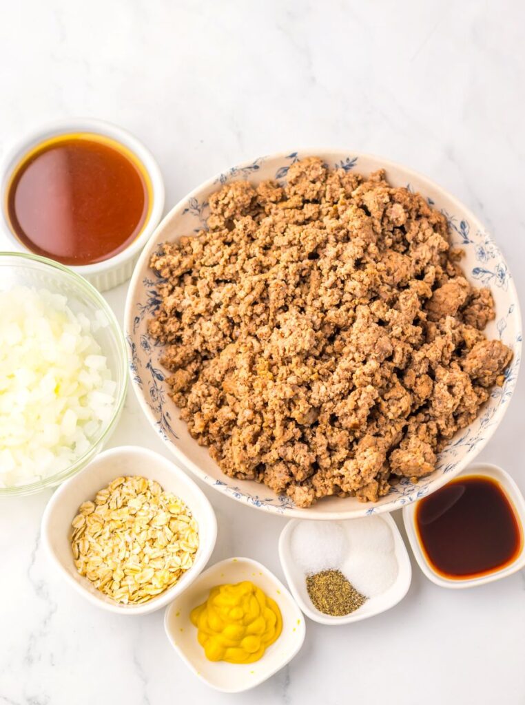 A bowl of cooked ground beef is surrounded by small bowls containing diced onions, oats, mustard, salt, pepper, barbecue sauce, and soy sauce, all arranged on a white marble surface.
