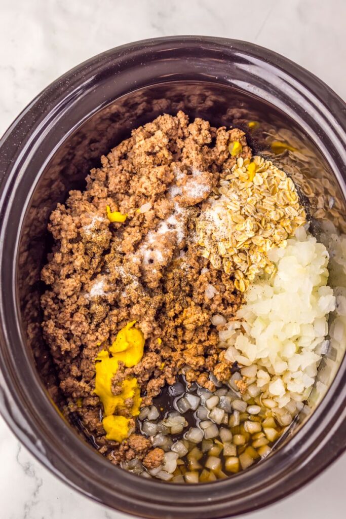 Ground beef, chopped onions, oats, mustard, and seasonings are layered in a slow cooker on a white marble surface, ready to be mixed for a recipe.