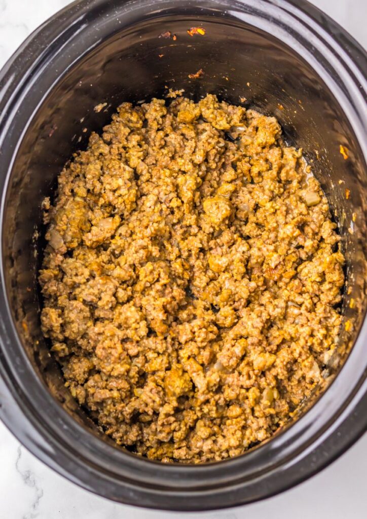 Ground beef with seasoning and small pieces of onion cooking in a black slow cooker, viewed from above. The mixture appears browned and well-mixed.