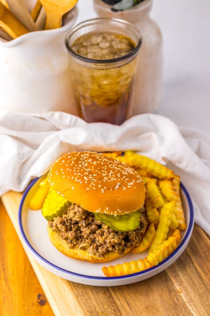 A plate with a Maid Rites sandwich topped with pickles and a serving of crinkle-cut fries, set on a wooden board. In the background, there’s a glass of iced drink and kitchen utensils in a white container.