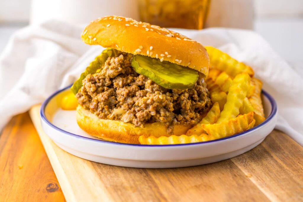 A Maid Rites sandwich topped with pickle slices on a sesame bun, served with crinkle-cut French fries on a white plate with a blue rim.