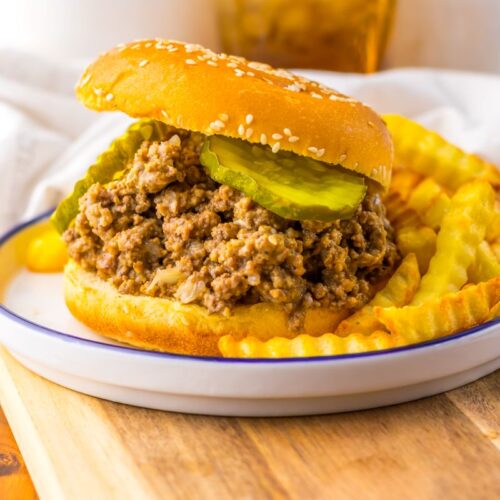 A Maid Rites sandwich topped with pickle slices on a sesame bun, served with crinkle-cut French fries on a white plate with a blue rim.