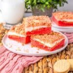 Three strawberry shortcake bars with crumb topping are stacked on a white plate, placed on a pink cloth with woven mat underneath. A glass of milk, green plant, and cookies are in the background.