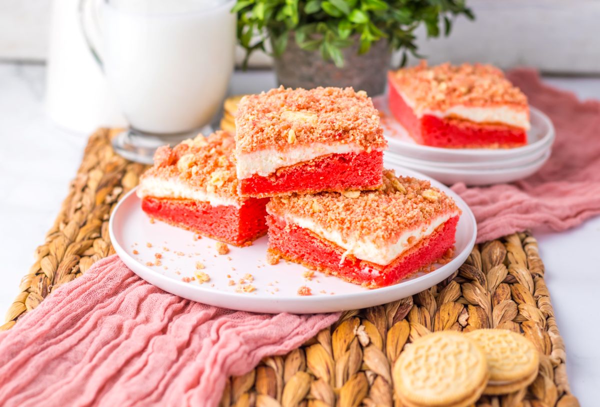 Three strawberry shortcake bars with crumb topping are stacked on a white plate, placed on a pink cloth with woven mat underneath. A glass of milk, green plant, and cookies are in the background.