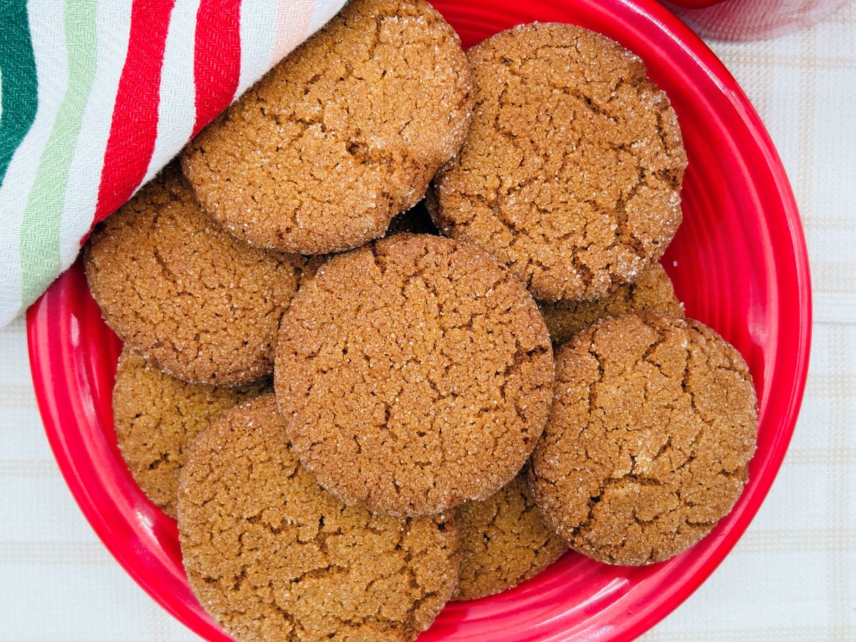 A red plate filled with golden-brown, sugar-coated cookies, partially covered by a striped red, green, and white kitchen towel on a light-colored surface.