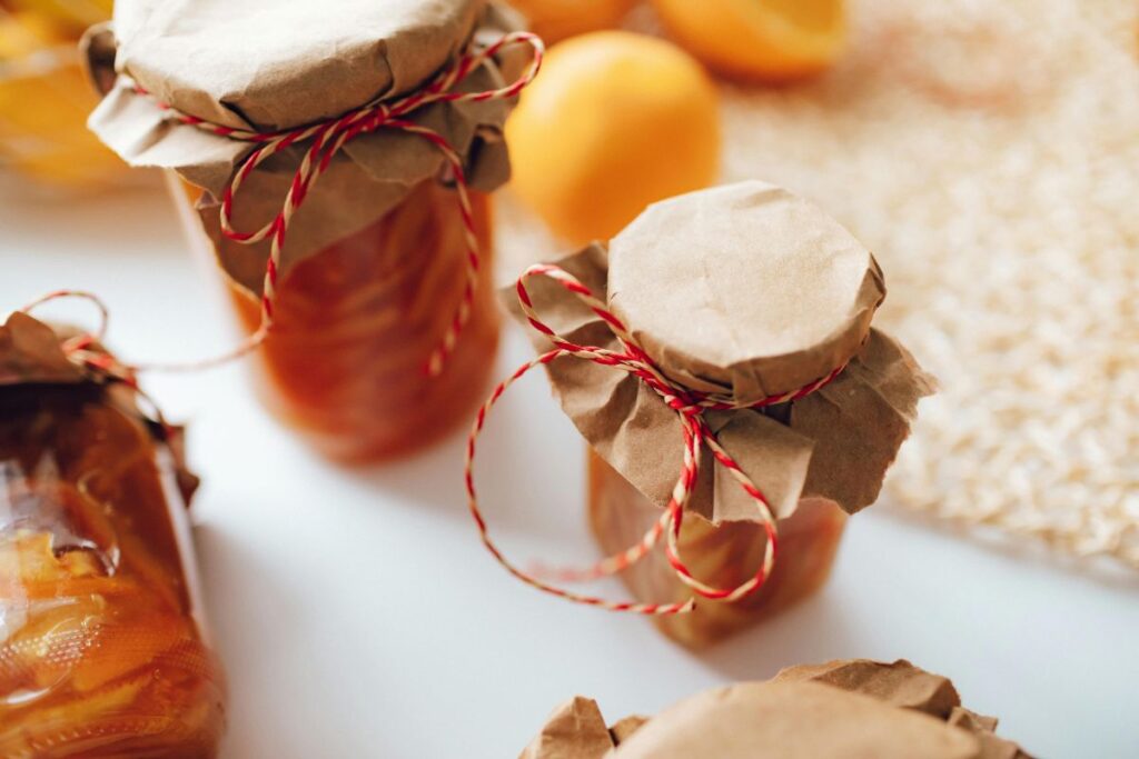 Glass jars filled with preserved fruit are covered with brown paper and tied with red and white string, sitting on a white surface with blurred fruit in the background.