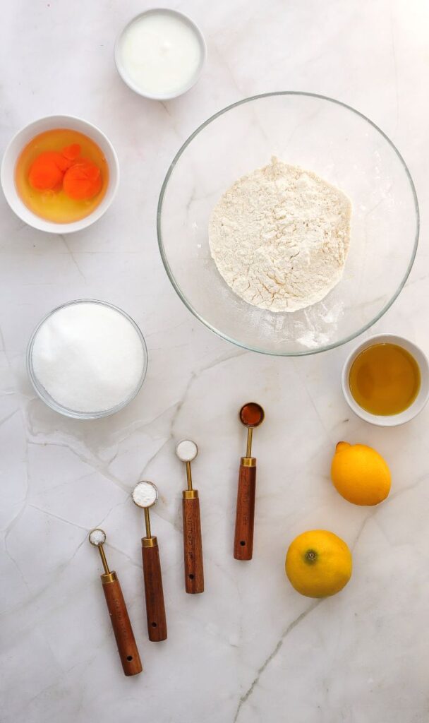 Baking ingredients on a marble surface: a bowl of flour, bowls with eggs, sugar, oil, and yogurt, two whole lemons, and four measuring spoons containing baking ingredients.