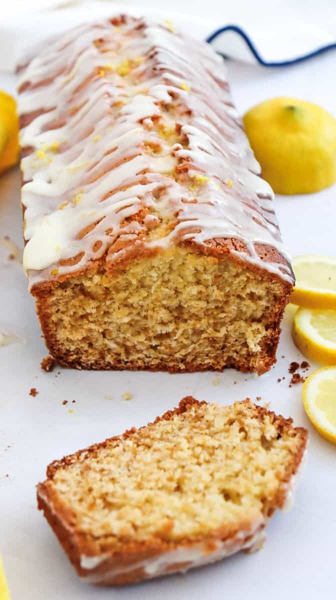 A loaf of lemon bread with a white glaze on top sits on a table next to lemon slices and whole lemons. One slice is cut and placed in front of the loaf, showing its moist, crumbly texture.
