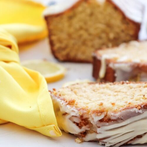 Two slices of glazed lemon pound cake sit on a white surface next to a yellow cloth, with a lemon and more cake in the background.