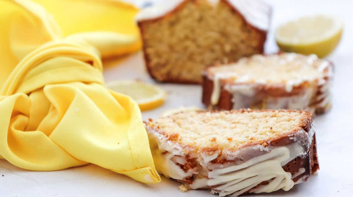 Two slices of glazed lemon pound cake sit on a white surface next to a yellow cloth, with a lemon and more cake in the background.