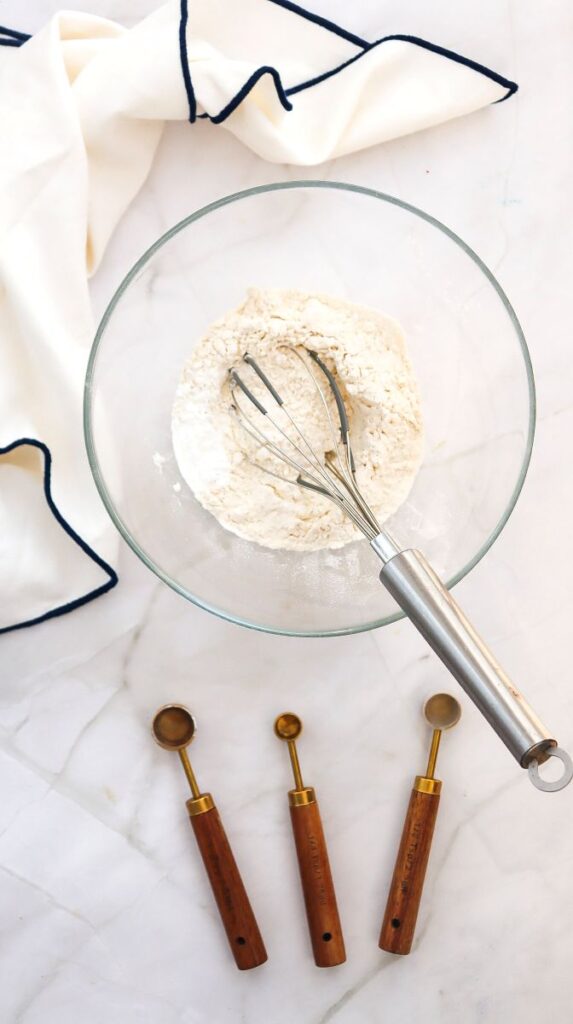 A glass bowl with flour and a whisk inside sits on a marble surface. Below the bowl are three wooden measuring spoons. A white cloth with a dark border is in the upper left corner.