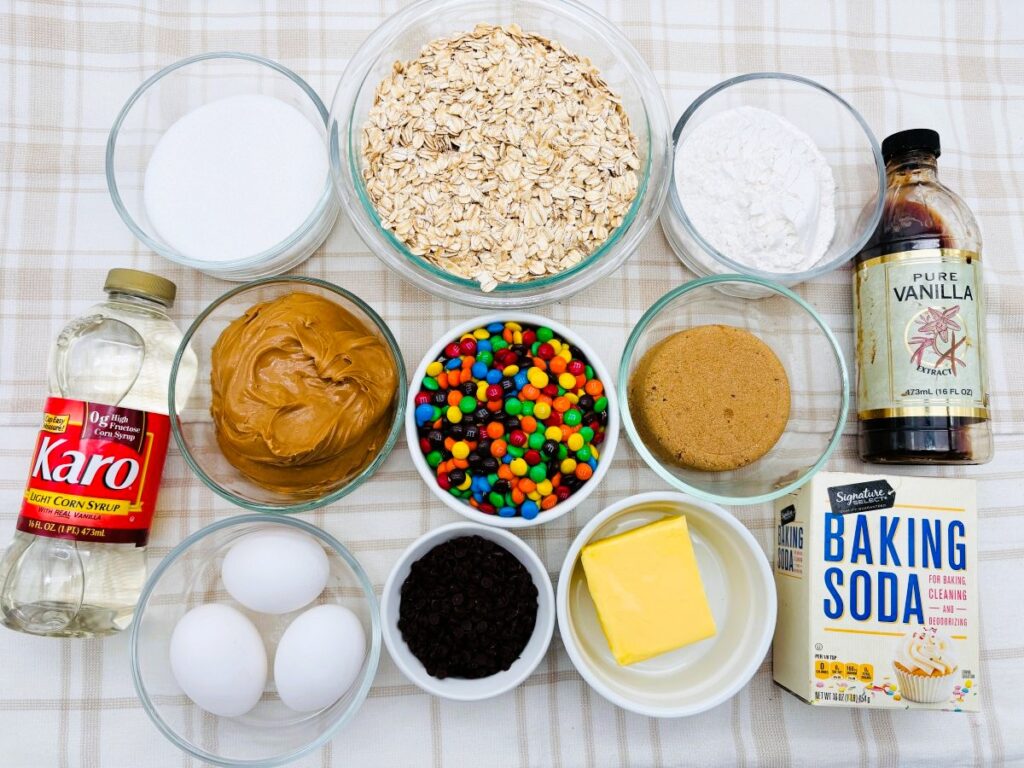 A top-down view of baking ingredients on a plaid tablecloth, including oats, sugar, flour, vanilla, corn syrup, peanut butter, brown sugar, MMs, eggs, butter, chocolate chips, and baking soda.