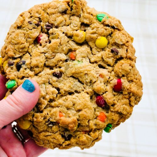 A hand with blue nail polish holds a large, round oatmeal cookie with colorful candy-coated chocolates and chocolate chips in it. The background is a light, plaid surface.
